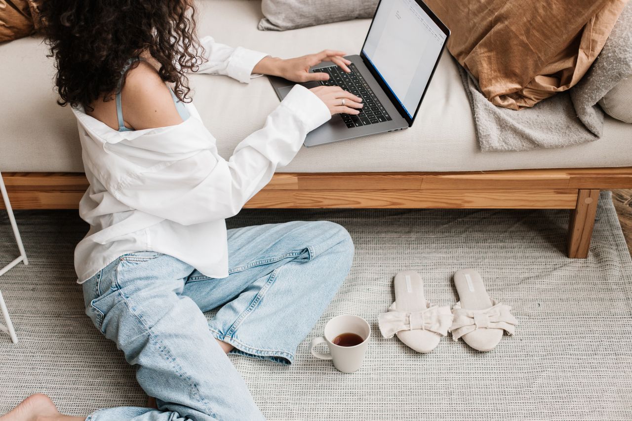 creative-03 Woman with curly hair typing on laptop while sitting on floor, surrounded by cozy elements.