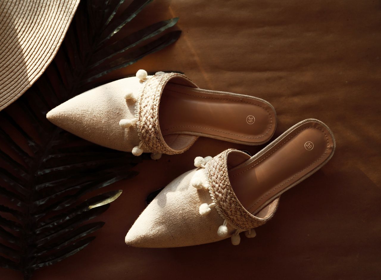 Services-01 Stylish beige sandals with decorative pom-poms on a summer backdrop, featuring woven straw elements.