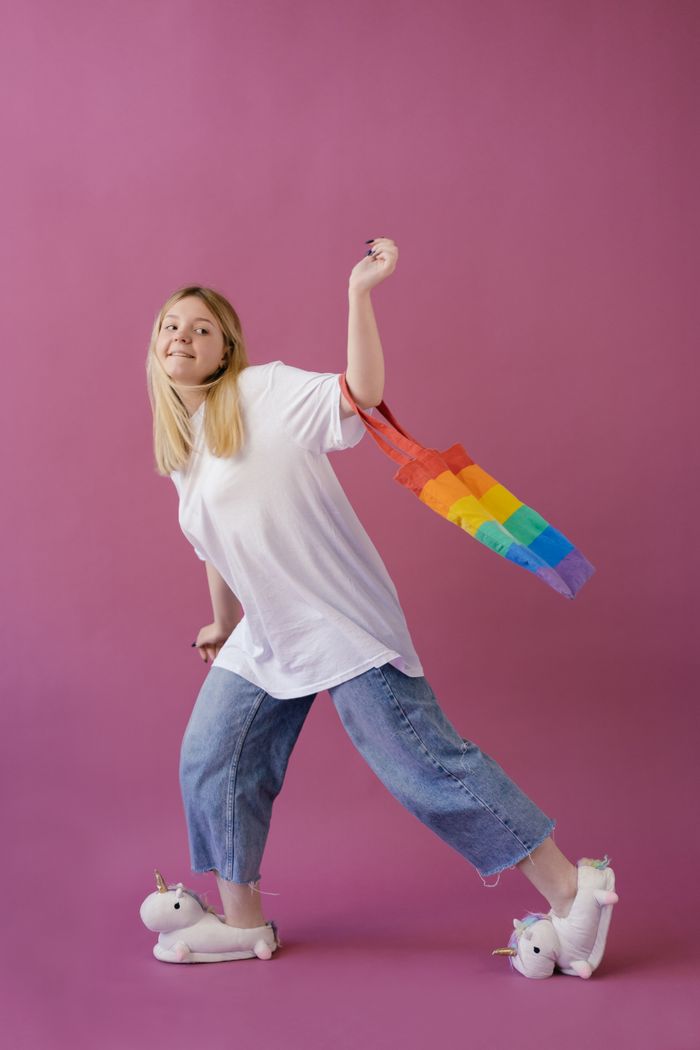 digital-01 A cheerful teenage girl posing with a rainbow tote bag, wearing comfy unicorn slippers on a pink backdrop.